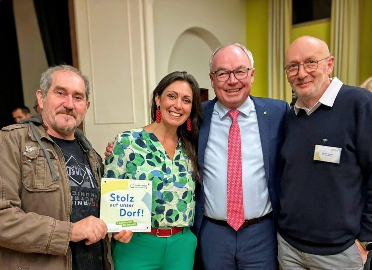 Vier Personen posieren für ein Foto. Eine Frau hält ein Schild mit der Aufschrift 'Stolz auf unser Dorf!' Ein Mann im Anzug mit roter Krawatte und zwei weitere Männer.