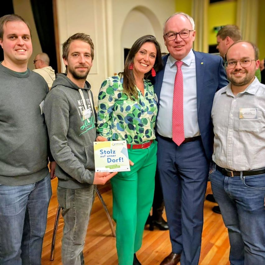 Fünf Menschen stehen lächelnd für ein Foto zusammen im Innenbereich. Ein Mann auf der linken Seite hält ein Buch mit dem Titel Stolz auf unser Dorf.