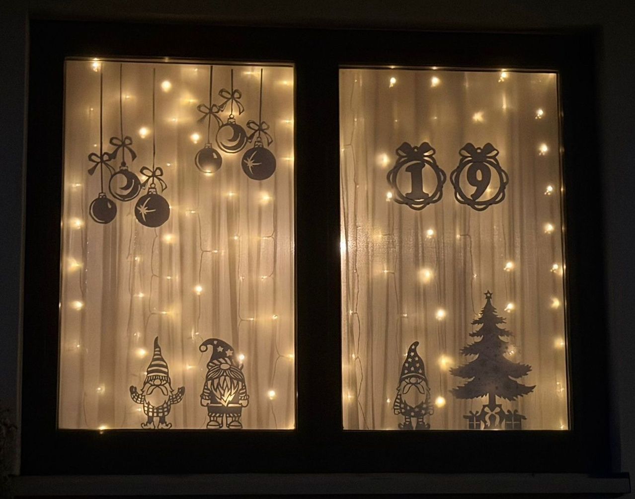 Eine Fensterdekoration zeigt hängende Weihnachtskugeln und Gnomen mit einem leuchtenden Weihnachtsbaum. Das Datum 19 ist über dem Baum angegeben.