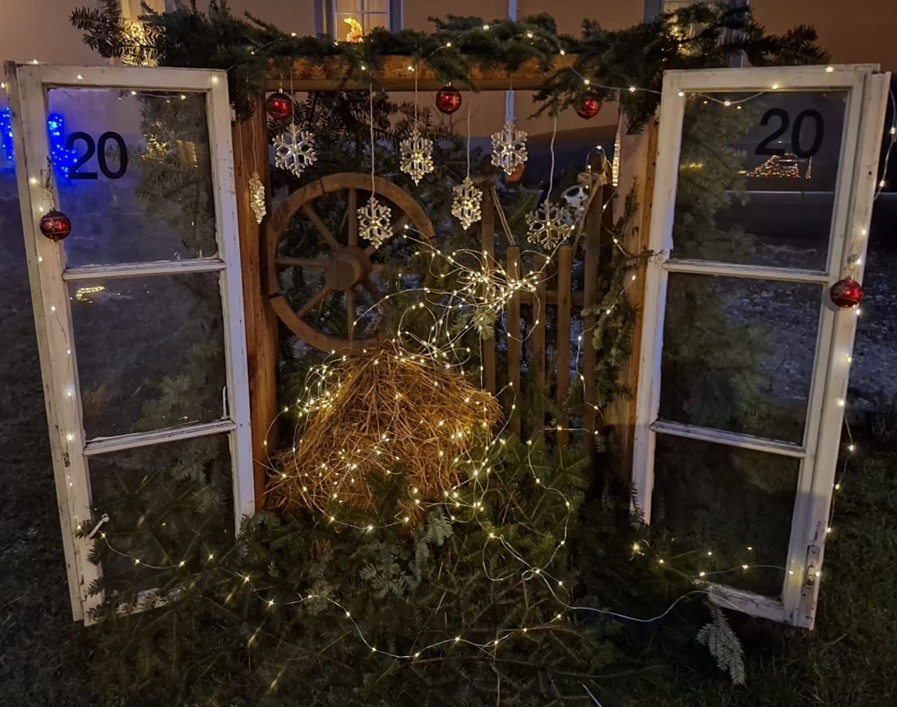 Eine Weihnachtsdekoration mit weißen Lichtern, einem Heuballen und Schneeflocken ist draußen ausgestellt. Die Fenster sind offen und es steht eine 20 auf einem von ihnen.