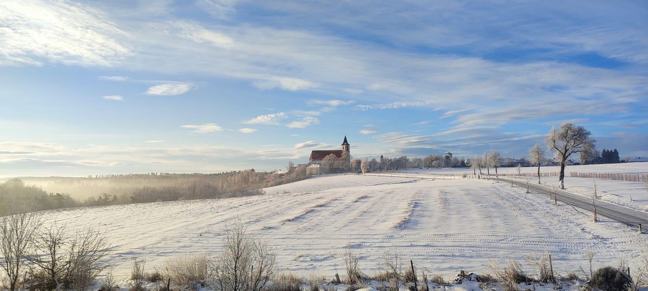 A snowy landscape with a church in the distance under a blue sky with scattered clouds.
