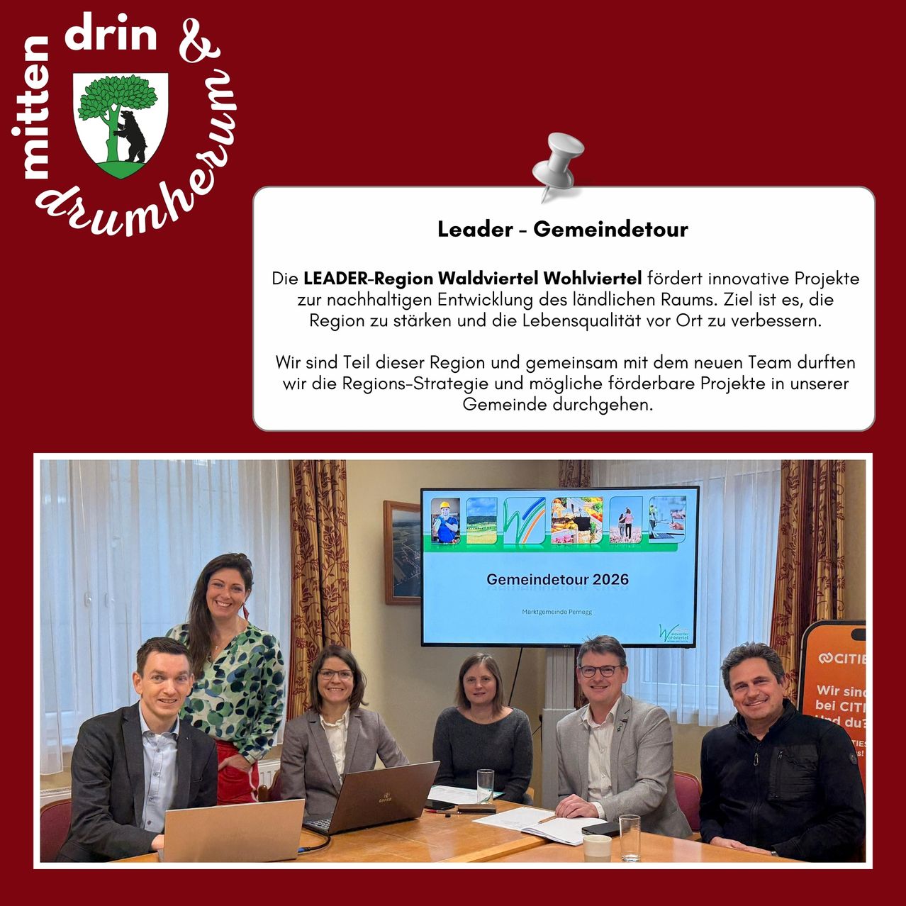 Gruppenfoto von sechs Personen, die um einen Tisch mit Laptops, Gläsern und Papieren sitzen. Ein Bildschirm hinter ihnen zeigt 'Gemeindetour 2026'. Der Hintergrund besteht aus einer weißen Wand mit Vorhängen und einem roten Rahmen.