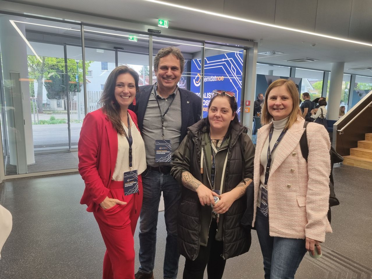 Vier Personen posieren für ein Foto in einem Flughafen. Ein Mann und drei Frauen, alle mit Namensschildern. Dahinter befindet sich ein großes blaues Banner mit weißen Text.