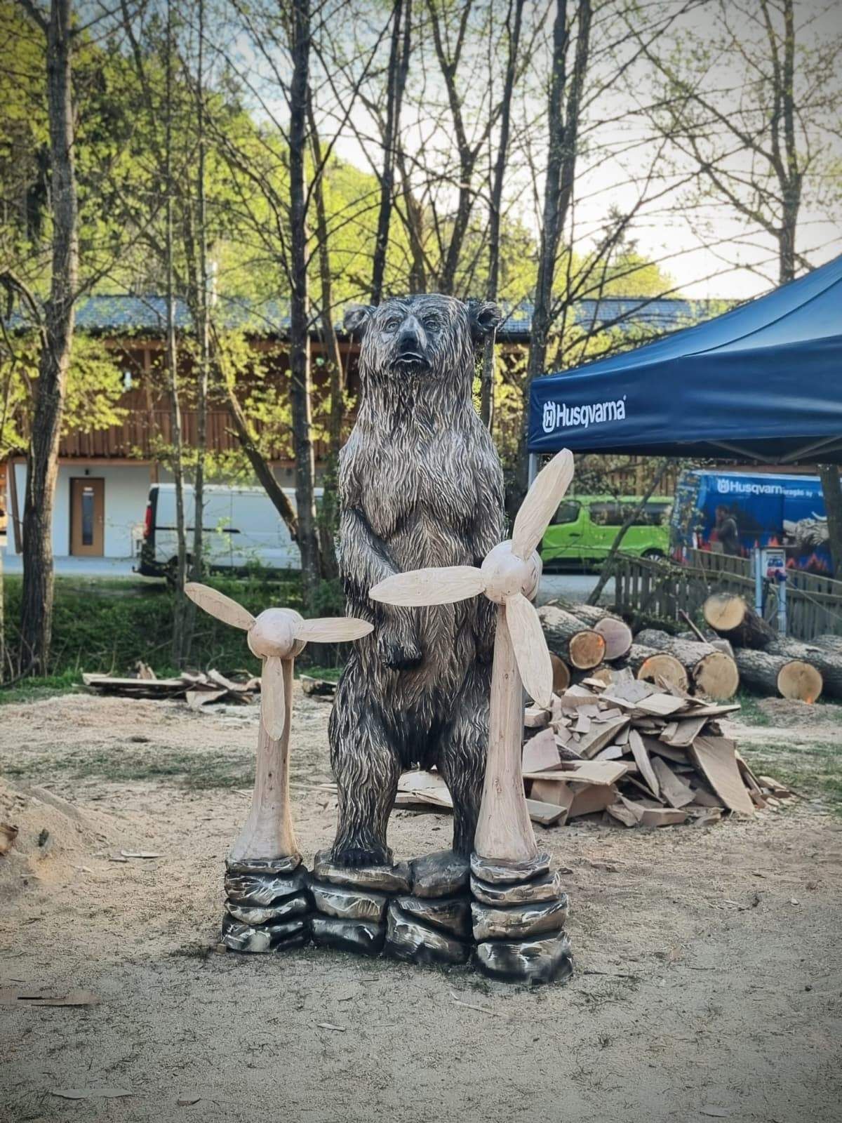 Ein geschnitzter Holzbär steht auf Felsen mit Windmühlen davor. Es befindet sich in einer Außenumgebung mit Bäumen, einem Gebäude und einem Zelt. Stapel von Holzscheiten sind in der Nähe.