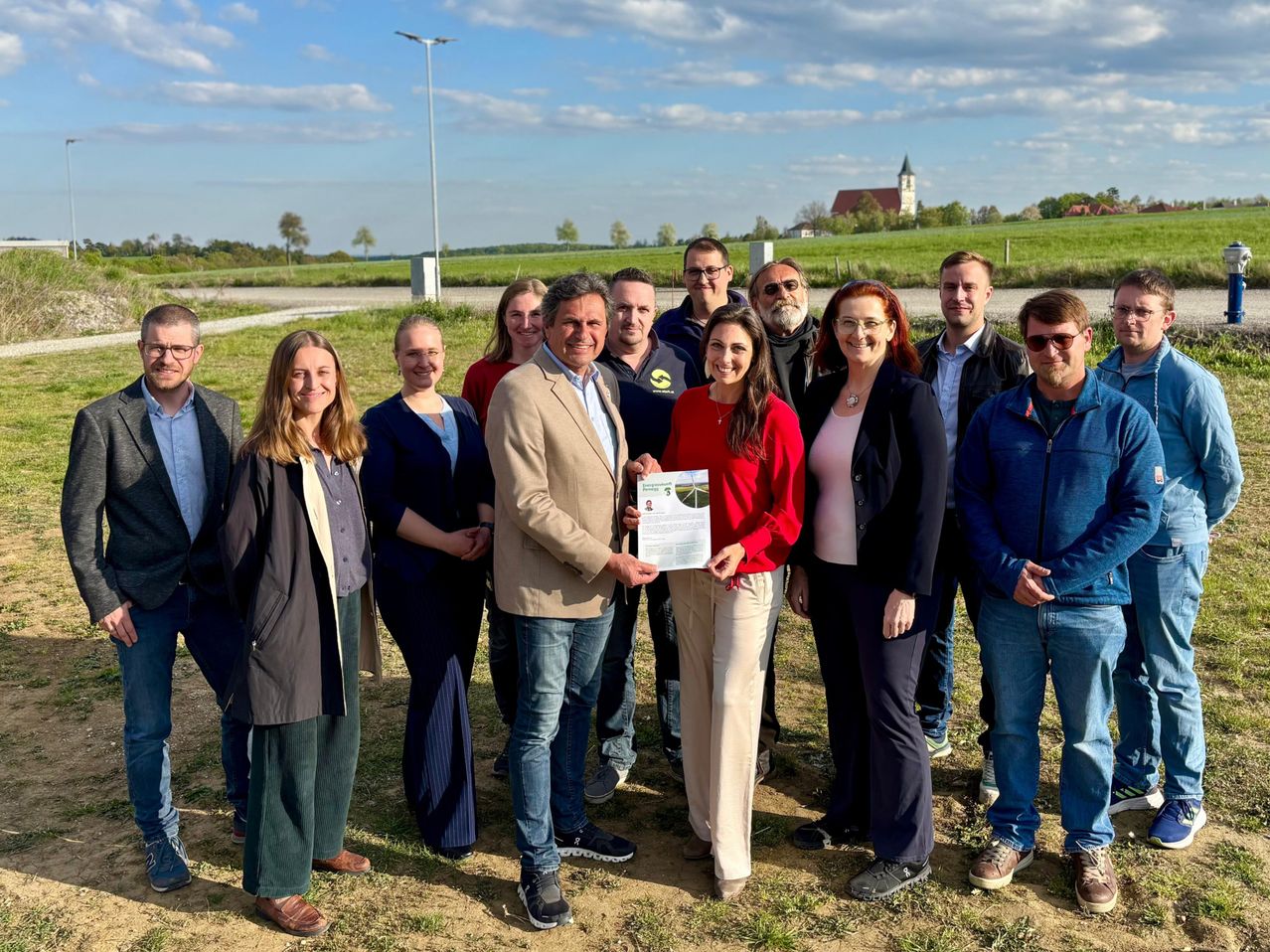 Eine Gruppe von Menschen steht auf einem Feld und hält ein Zertifikat. Sie sind in Freizeitkleidung gekleidet und lächeln für ein Foto. Im Hintergrund ist eine ländliche Landschaft mit Feldern und einer Kirche zu sehen.