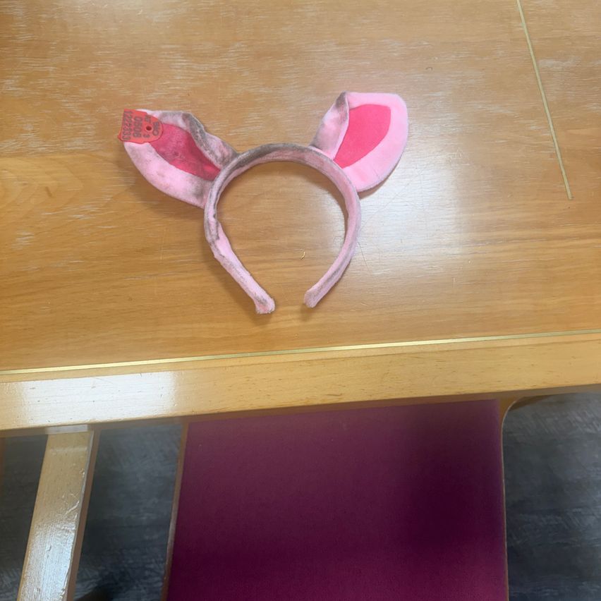 Ein rosa Schwein-Kopfband liegt auf einem Holztisch. Das Kopfband hat eine rosa Farbe und einen Anhänger. Der Tisch hat eine hölzerne Textur.