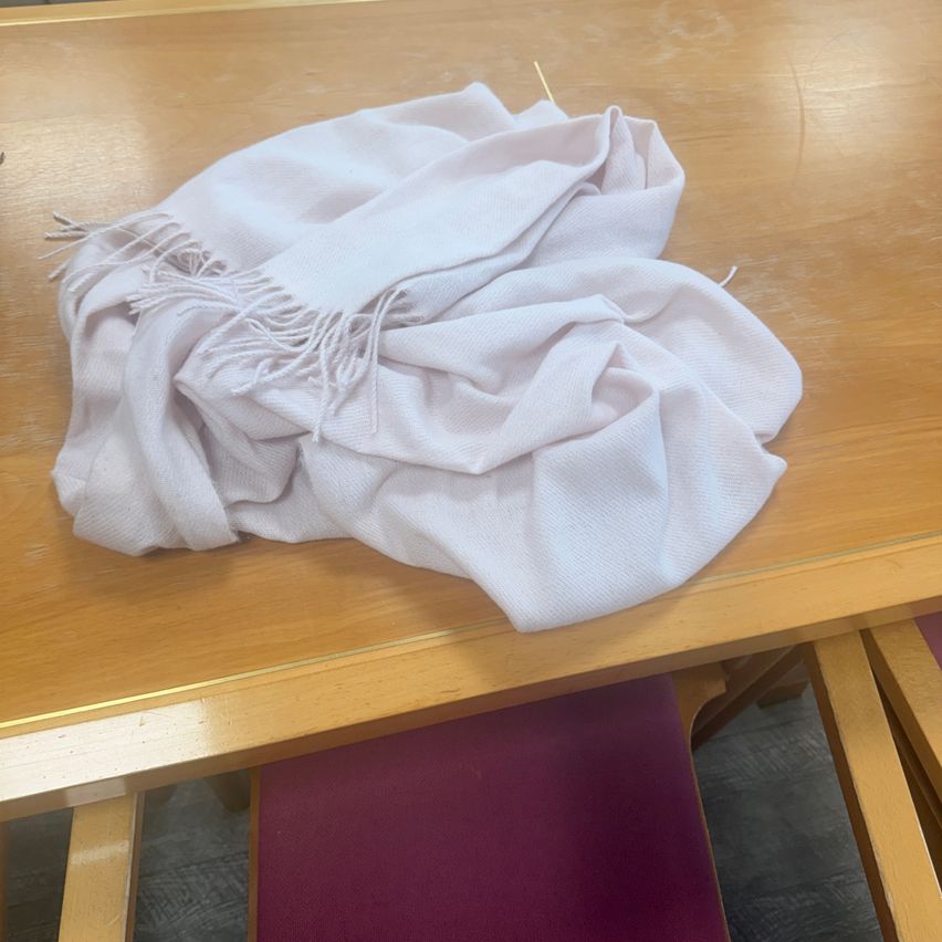 Ein hellrosa Schal mit Fransen liegt auf einem hölzernen Tisch mit glänzender Oberfläche. Ein rot gepolsterter Stuhl ist neben dem Tisch zu sehen.