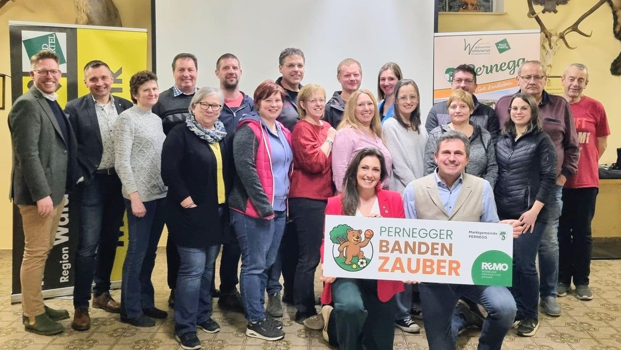 Eine Gruppe Erwachsener in Freizeitkleidung posiert für ein Foto in einer Innenumgebung, wobei eine Frau ein Schild mit der Aufschrift 'Pernegger Banden Zauber' hält.