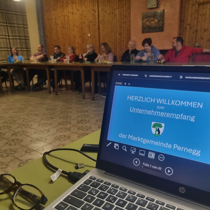 Ein Laptop mit geöffnetem Bildschirm, der eine Willkommensnachricht zu einem Unternehmenstreffen zeigt, mit Menschen, die am Tisch im Hintergrund sitzen.