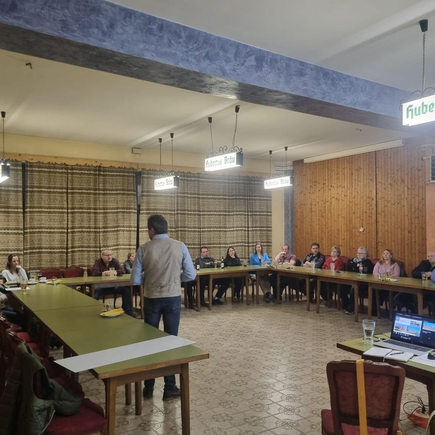 Ein Mann steht an einem Tisch in einem Konferenzraum und spricht zu Personen, die an langen Tischen sitzen. Der Raum hat Holzwände und Decken, mit hängenden Lampen und Schildern. Die Tische sind mit Tellern, Gläsern und Laptops bestückt.