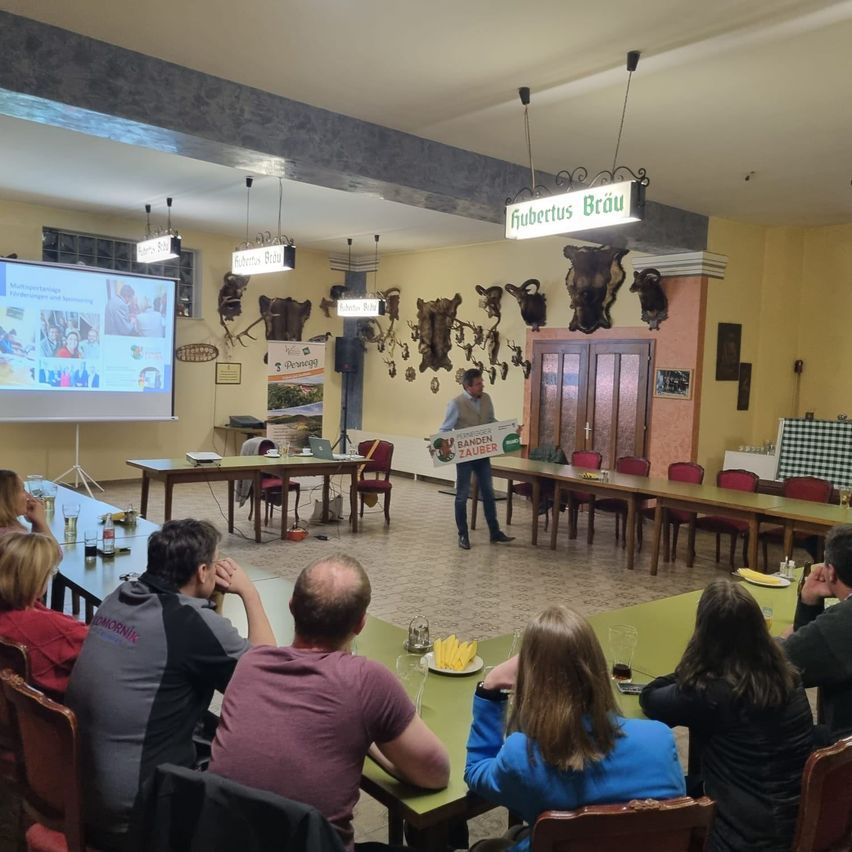 In einem Speisesaal steht ein Mann vor einer Projektionsleinwand und zeigt eine Präsentation. Mehrere Personen sitzen um einen langen Tisch herum und lauschen. Der Raum hat Tierköpfe und Lampen, die von der Decke hängen.