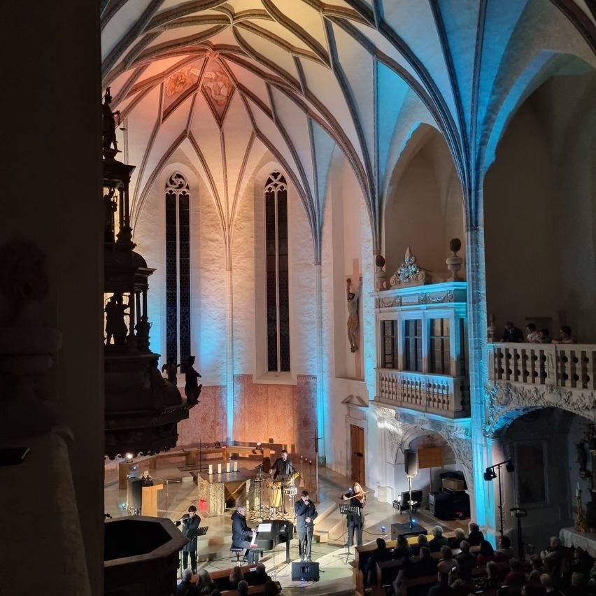 Eine Aufführung in einer Kathedrale mit Musikern auf der Bühne, einem Publikum in den Bänken und dekorativen Elementen an den Wänden und der Decke.