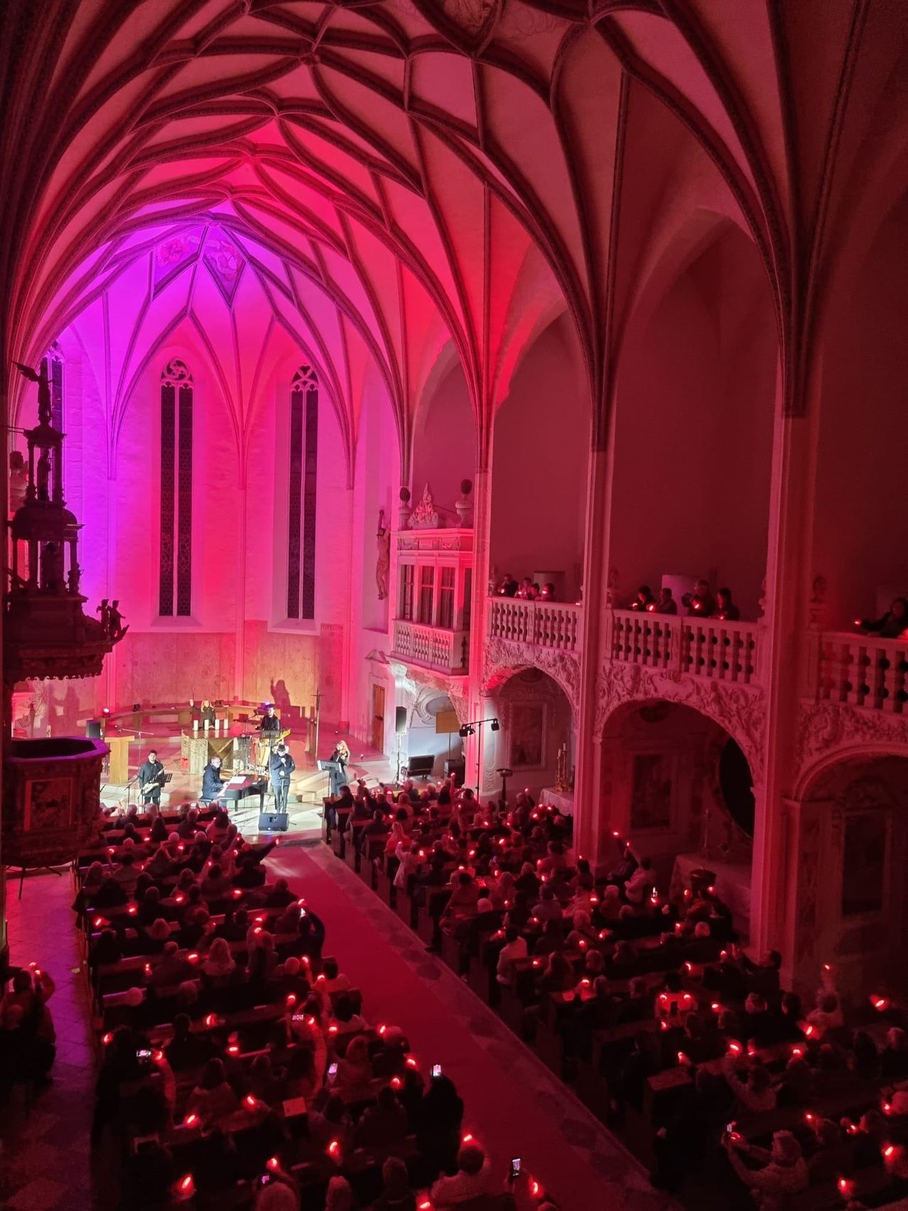 Eine schwach beleuchtete Kirche mit Bühne, Musikern und einer Menge mit Kerzen. Die Bühne ist mit rosa Lichtern beleuchtet.
