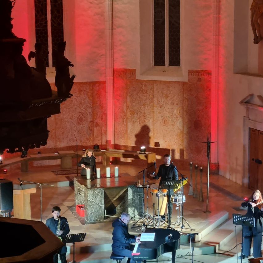Eine Band spielt in einer Kirche mit roter Beleuchtung. Eine Person spielt Klavier, eine andere Geige, eine dritte Schlagzeug und die vierte Saxophon.