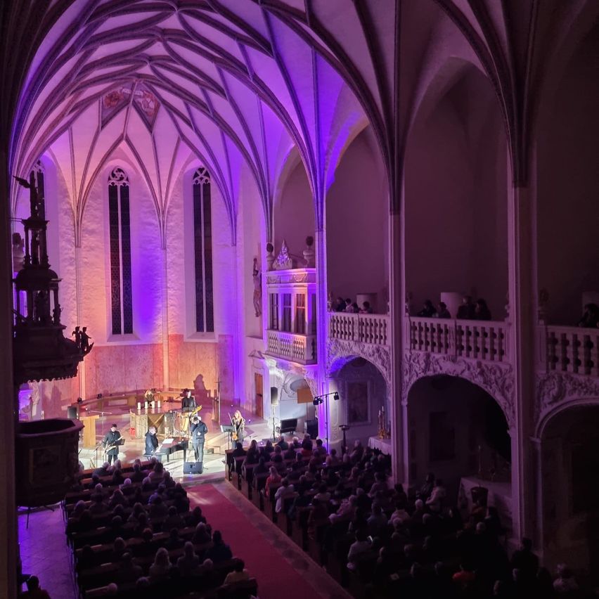 Eine Band spielt auf einer Bühne in einer Kathedrale mit lila Beleuchtung. Ein Publikum sitzt unten und Menschen sind auf der Galerie.
