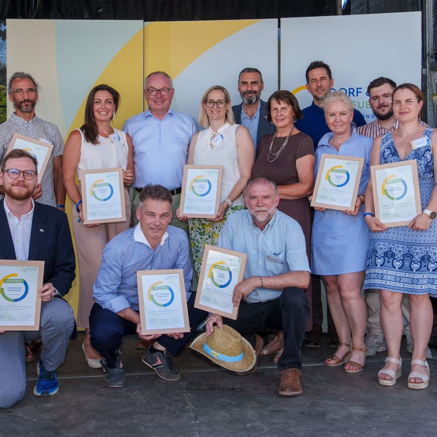 Eine Gruppe von Menschen, sowohl Männer als auch Frauen, posiert für ein Foto, während sie Urkunden halten und lächeln. Sie stehen vor einem Hintergrund mit einem gelben und weißen Design.