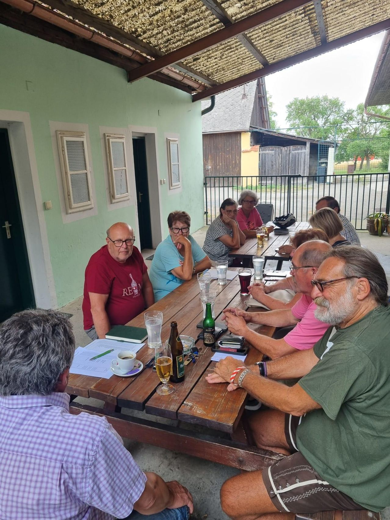 Eine Gruppe von Menschen sitzt um einen Holztisch im Freien, einige mit Brillen und Büchern, eine Tasse und eine Flasche Bier.