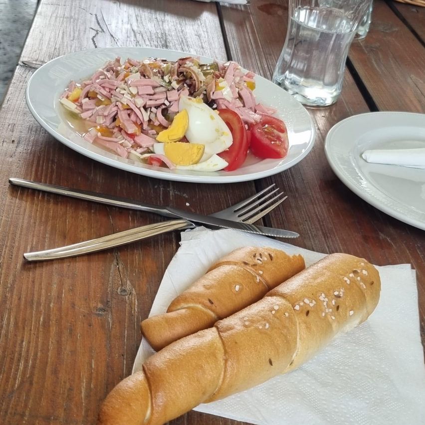 Ein Holztisch zeigt einen Teller mit gemischtem Salat mit Schinken, Eiern und Tomaten, begleitet von zwei Brötchen auf einem Serviettentuch, einer Gabel und einem Messer.