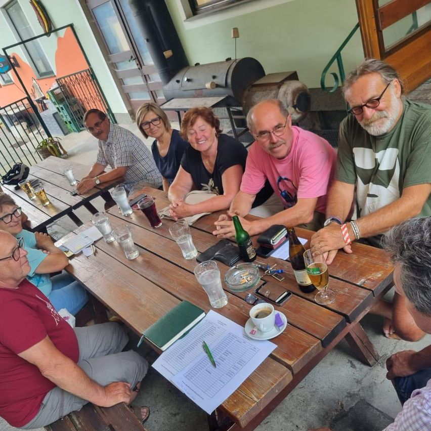 Eine Gruppe von Menschen sitzt draußen an einem Holztisch, mit Gläsern, einem Notizbuch und einem Stift auf dem Tisch. Sie lächeln und scheinen in ein Gespräch vertieft zu sein. Im Hintergrund befinden sich ein Metallgeländer, ein Grill und ein Gebäude.