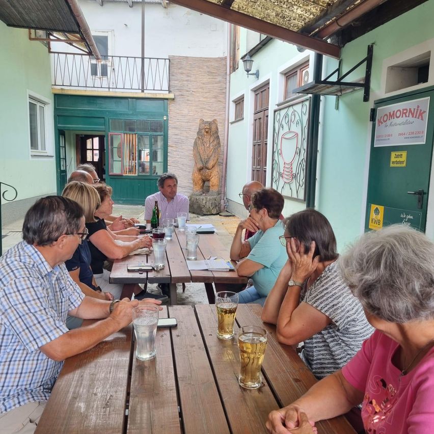 Eine Gruppe von Menschen sitzt um einen Holztisch in einem Restaurant im Freien, einige tragen Brillen und halten Biergläser. Ein Mann steht neben einem Tisch mit einer Flasche und Papieren. Eine Bärenstatue steht dahinter.