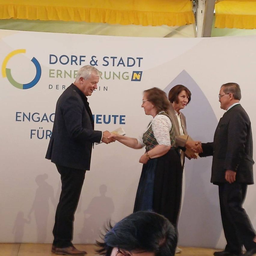 Vier Personen stehen vor einem Banner, zwei Männer und zwei Frauen, und schütteln Hände. Sie scheinen bei einer Veranstaltung zu sein.