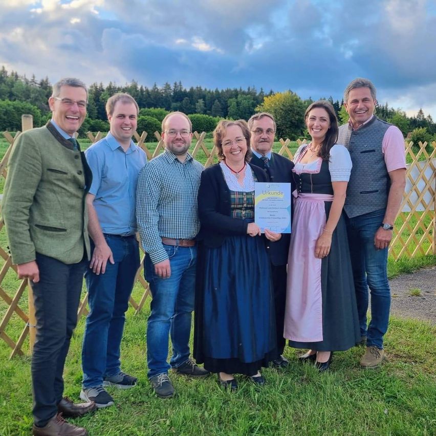 Eine Gruppe von Menschen steht auf einem Rasenfeld mit einem Zaun im Hintergrund. Sie halten eine Urkunde und sehen glücklich aus. Bäume und ein bewölkter Himmel sind hinter ihnen.