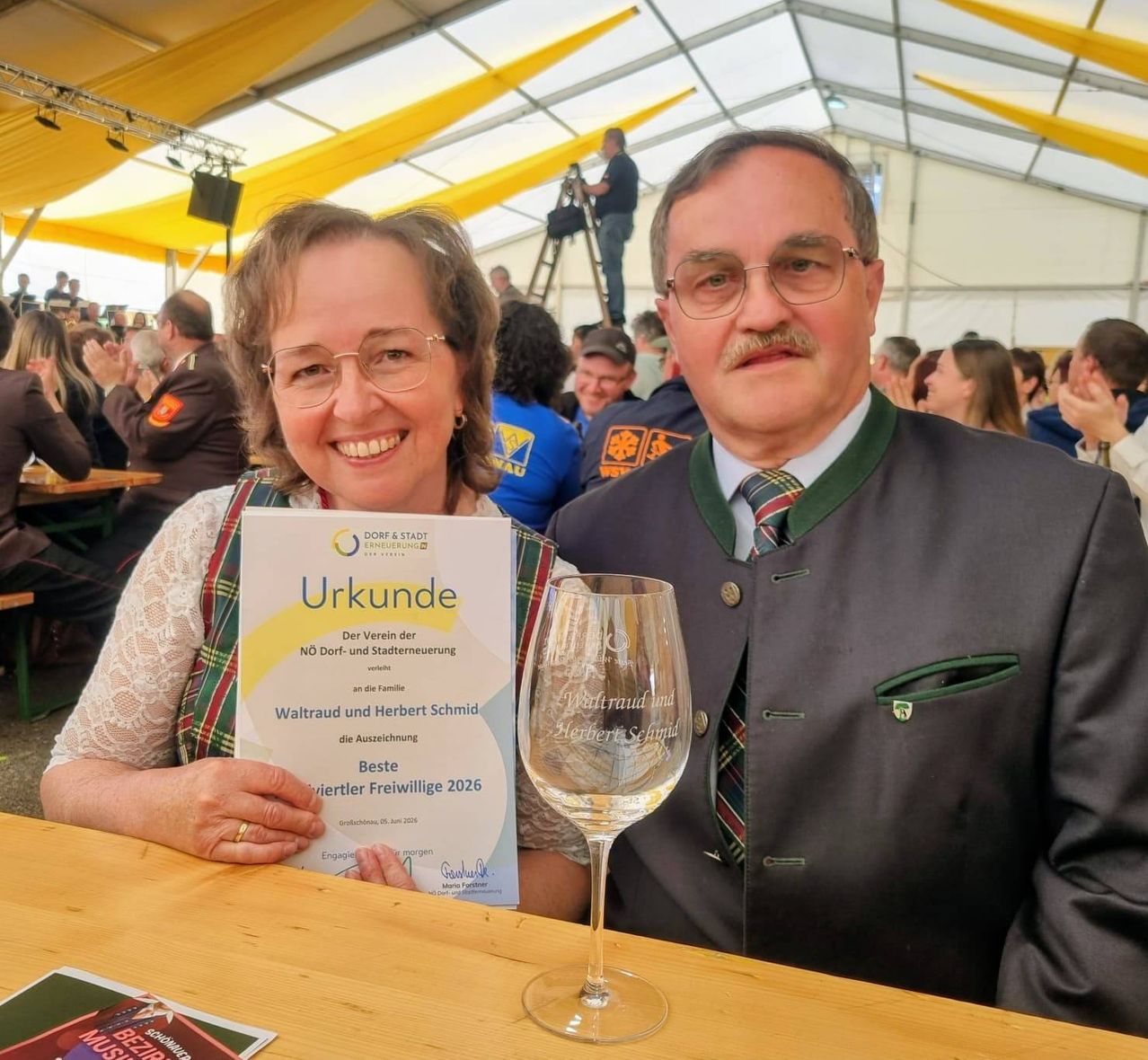 Eine lächelnde Frau und ein Mann posieren für ein Foto auf einem Festival. Sie halten ein Zertifikat und ein Weinglas. Die Frau trägt eine Brille und einen Ring. Der Mann trägt einen Anzug und eine Brille. Sie sind von Menschen umgeben. Das Zelt hat ein gelb-weißes Dach.