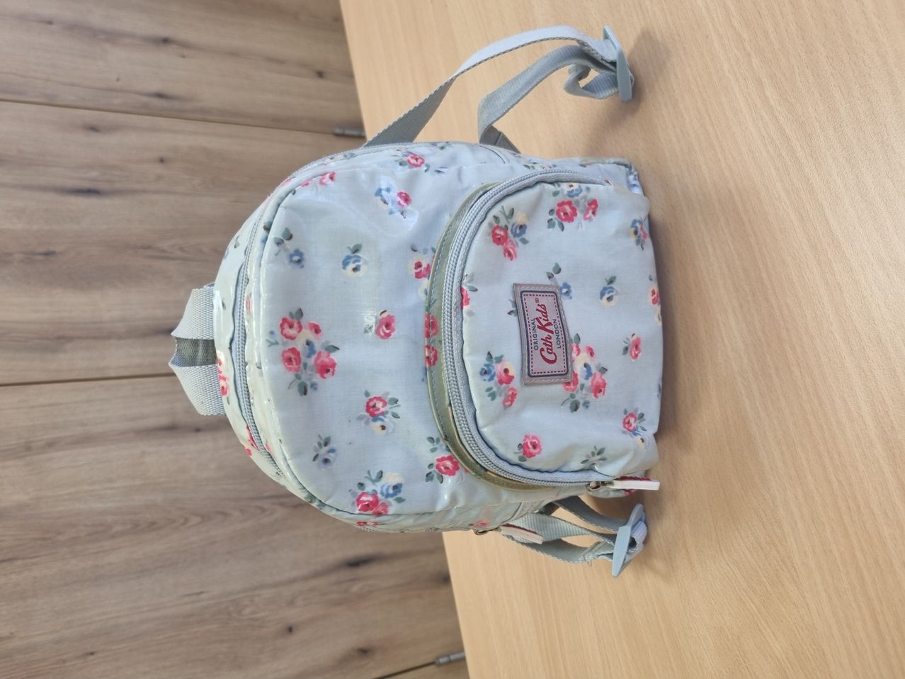 Ein hellblauer Rucksack mit Blumenmuster auf einer Holzoberfläche. Er hat mehrere Reißverschlüsse und verstellbare Träger.