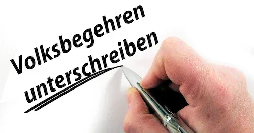 Eine Hand hält einen Stift und schreibt auf ein weißes Blatt mit den Worten 'Volksbegehen unterschreiben'. Die Hand ist teilweise sichtbar.