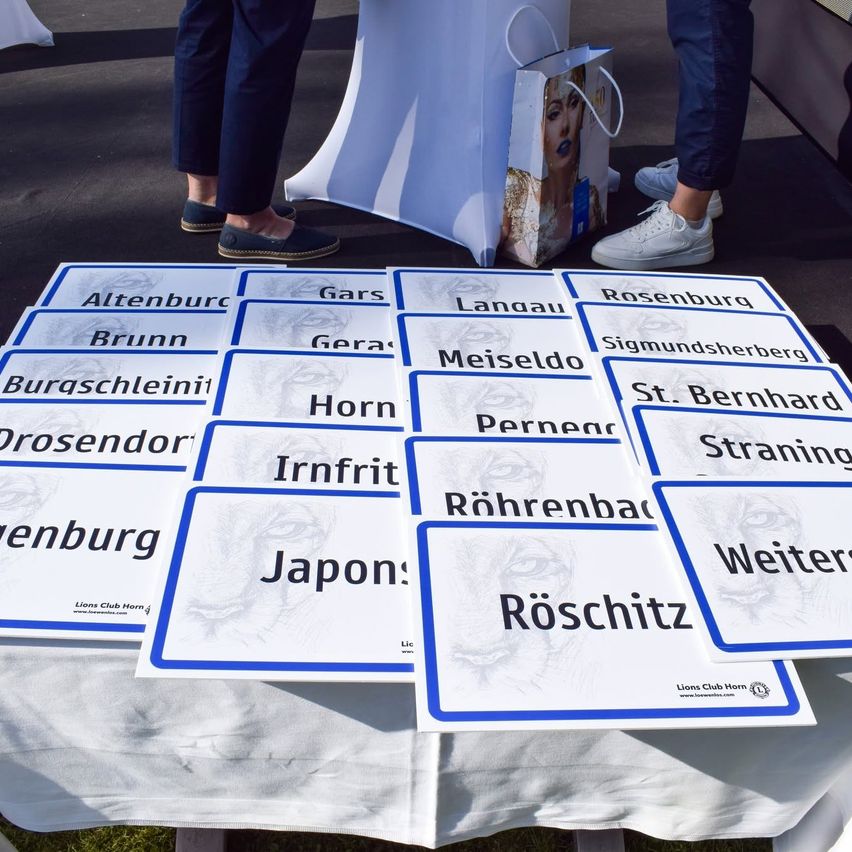 A table displays blue and white cards with the names of different places. Two people stand nearby, one in white sneakers, the other in loafers. A paper bag with a woman's image is on the table.