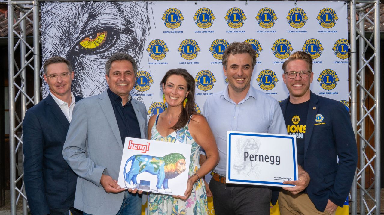Four individuals, smiling, pose with lion-themed items. A man holds a lion statue, and a woman holds a sign with a lion's head. The backdrop features multiple Lions Club logos.