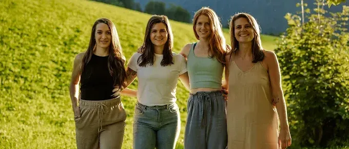 Vier Frauen posieren an einem sonnigen Tag auf einer Wiese, mit Bergen im Hintergrund.