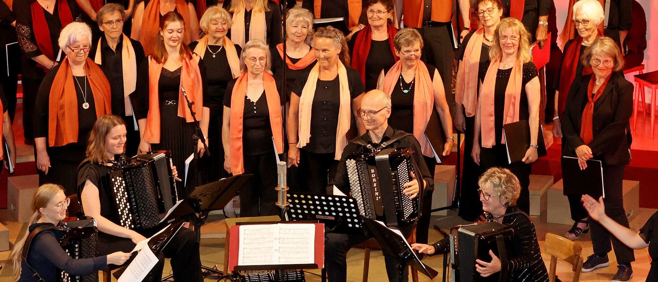 Ein Chor in Schwarz und Orange versammelt sich auf der Bühne und tritt mit Musikinstrumenten auf. Sie werden von einem Publikum begleitet.