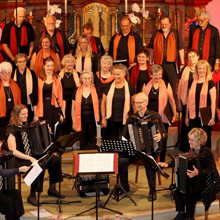 Ein Chor in schwarz und orange tritt auf der Bühne auf, spielt Musikinstrumente, darunter Akkordeons, und einige Leute stehen hinter ihnen und halten Notenblätter.