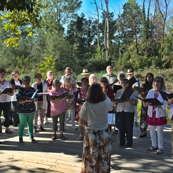 Eine Gruppe von Menschen in Hüten und Brillen singt auf einer Holzterrasse in einem Wald.