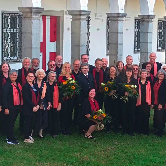 Eine Gruppe von Menschen in Schwarz und Rot mit Blumensträußen posiert für ein Foto vor einem Gebäude mit Säulen und einer roten Flagge.