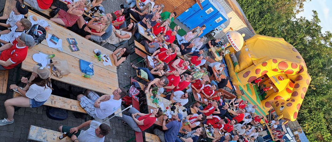 Viele Menschen sitzen an Picknicktischen im Freien, tragen rote Hemden. Sie essen und trinken. In der Nähe befinden sich blaue mobile Toiletten, ein aufblasbares Haus und Bäume.