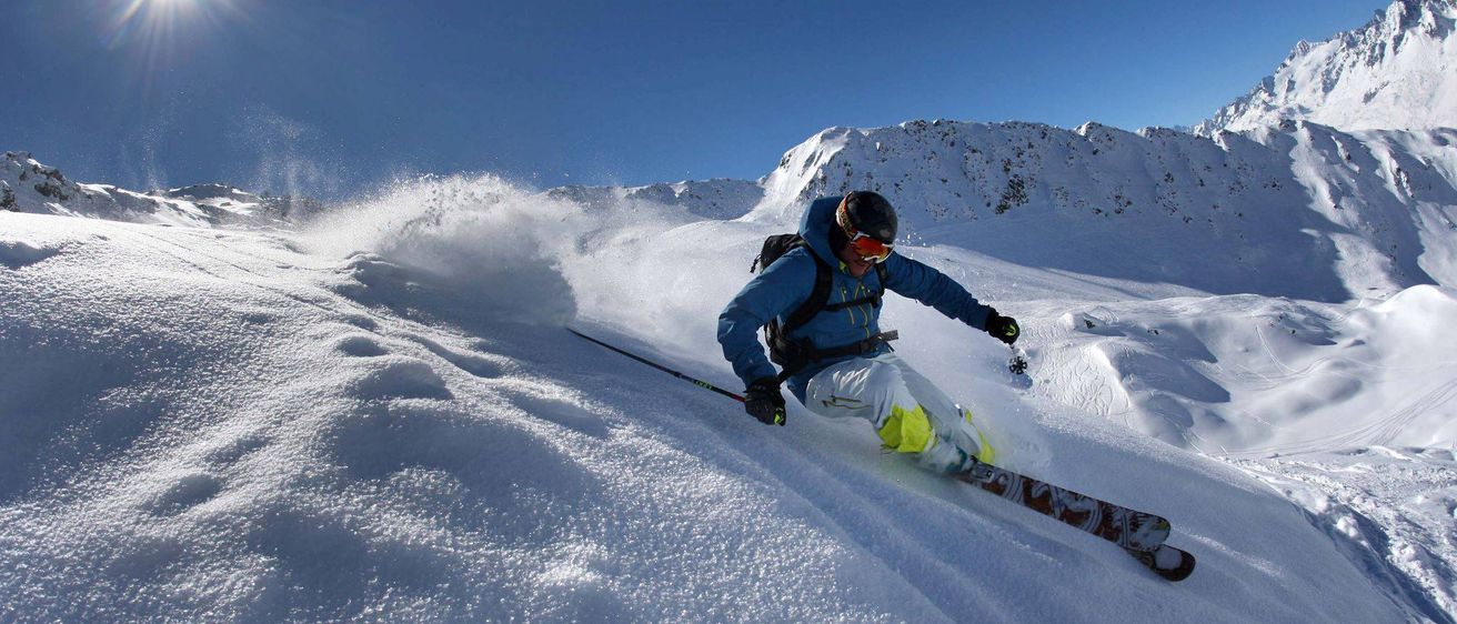 Ein Skifahrer in einer blauen Jacke und Helm fährt einen verschneiten Berg hinunter, mit fliegendem Schnee um ihn herum.