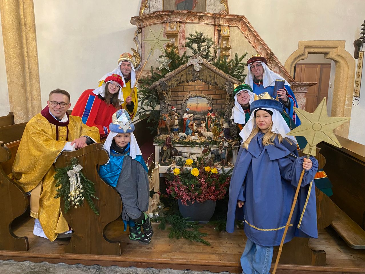 Eine Gruppe von Erwachsenen und Kindern in Weihnachtskostümen posiert vor einem Kirchenaltar mit einer Krippe und Blumen.