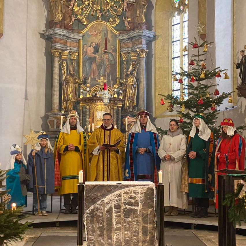 In einer Kirche singen eine Gruppe von Menschen in traditioneller Kleidung ein Weihnachtslied. Dahinter steht ein Weihnachtsbaum und ein goldener Altar.
