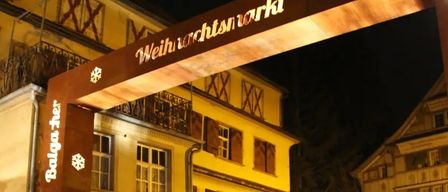 Illuminated arch at night over a building entrance, with the word 'Weihnachtsmarkt' written on it, featuring snowflakes and the word 'Bolgacher'.
