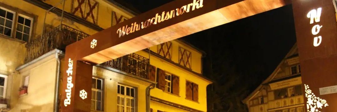 Beleuchteter Bogen in der Nacht über einem Gebäudeeingang, mit dem Wort 'Weihnachtsmarkt' darauf, mit Schneeflocken und dem Wort 'Bolgacher'.