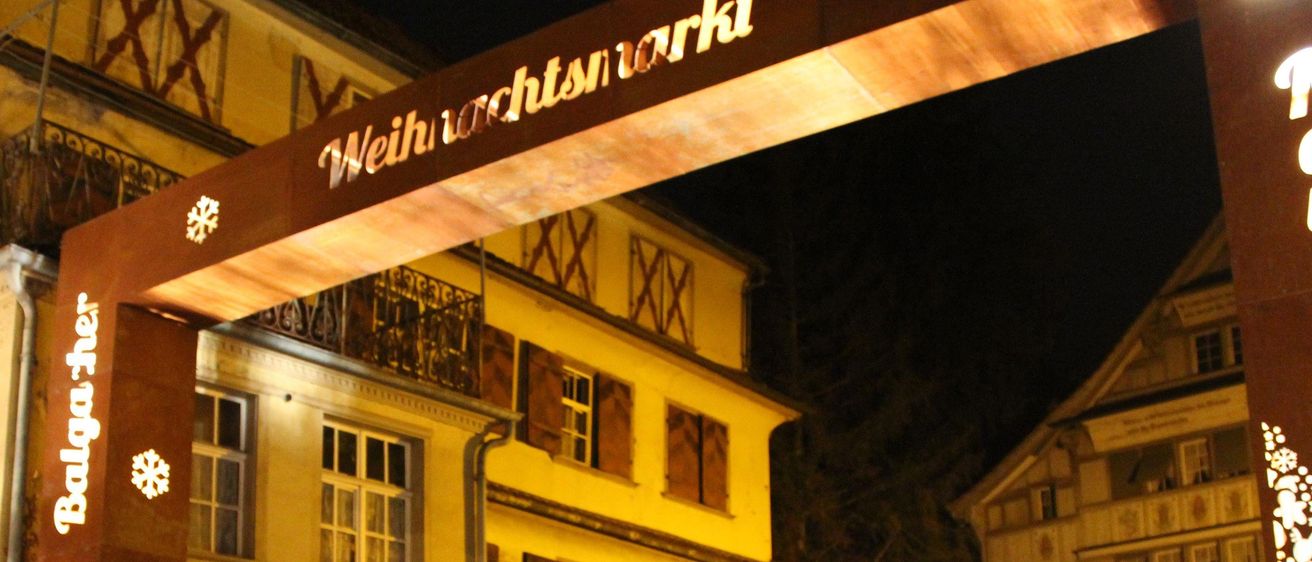 Ein Gebäude mit Balkon und Fenstern ist nachts beleuchtet. Ein beleuchtetes Schild darüber steht 'Weihnachtsmarkt'.
