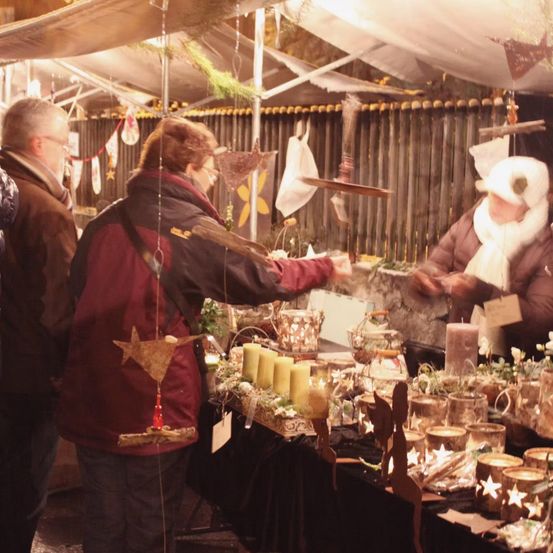 A market stall with candles and holiday items is under a canopy. Two people are looking at the items on the table.