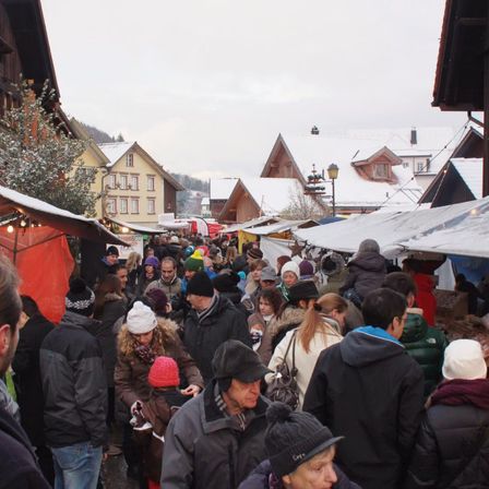 Eine belebte Marktstraße in einer verschneiten Stadt. Menschen in Winterkleidung navigieren durch den überfüllten Bereich, mit Zelten und Gebäuden in der Umgebung.