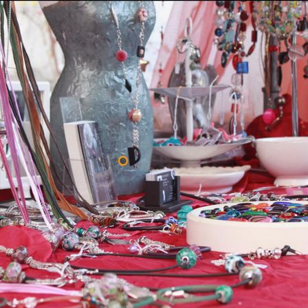 Ein mit rotem Stoff bedeckter Tisch zeigt verschiedene Halsketten und Armbänder, einschließlich Perlenketten und Silberketten. Im Hintergrund steht ein Mannequin, das Silberstoff trägt und mit einer roten und schwarzen Halskette geschmückt ist.
