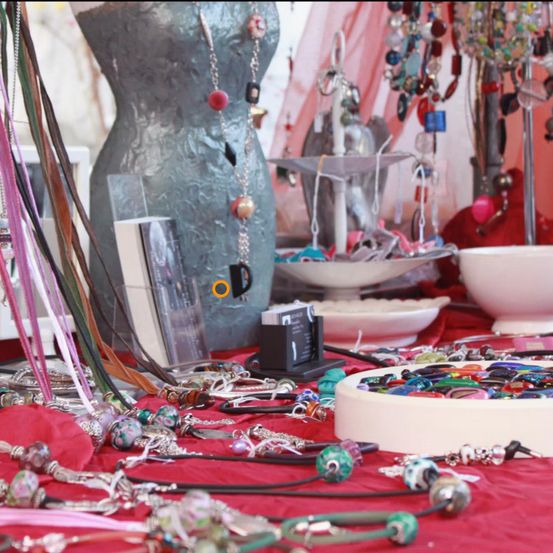 A table covered with red fabric displays various necklaces and bracelets, including beaded and silver chains. A mannequin wearing silver fabric and adorned with a red and black necklace is in the background.