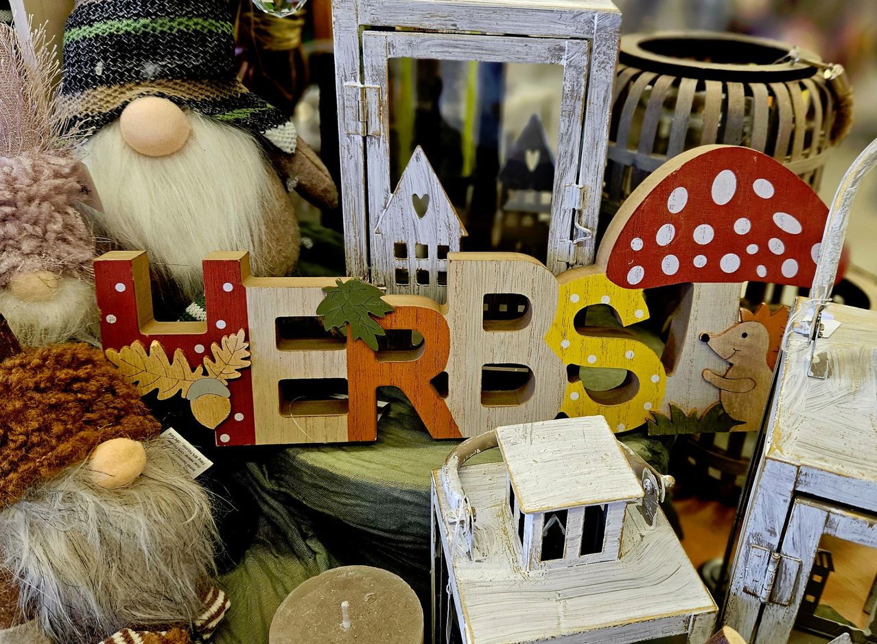Eine Anordnung von Holzbuchstaben buchstabiert 'Herbst' mit verschiedenen Herbstdekorationen wie einem Pilz, einem Gnom und einem Haus, platziert auf einer grünen Oberfläche.