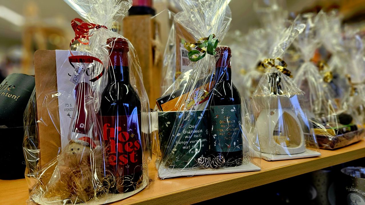 Weinflaschen, in Plastik eingewickelt und mit Geschenkbändern, sind auf einem Tisch ausgestellt. Ein Becher mit Weihnachtsthema und eine kleine Tannenzapfen sind neben den Flaschen platziert. Die Weinflaschen haben Etiketten mit Text und Illustrationen. Andere Gegenstände sind ebenfalls auf dem Tisch, in Plastik verpackt.