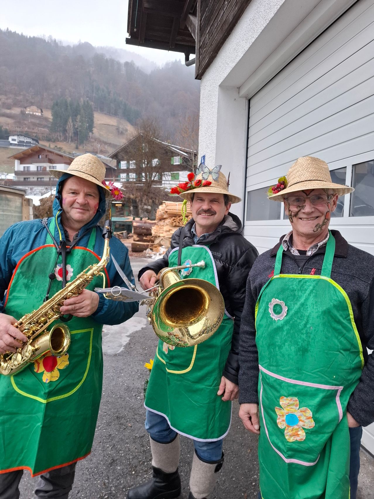 Drei Männer in grünen Schürzen und Strohhüten stehen vor einem Gebäude, einer hält eine Saxophon und der andere eine Trompete.